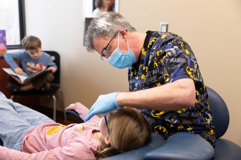 Dentist in blue scrubs working on a patient in a dental chair; bright office setting.